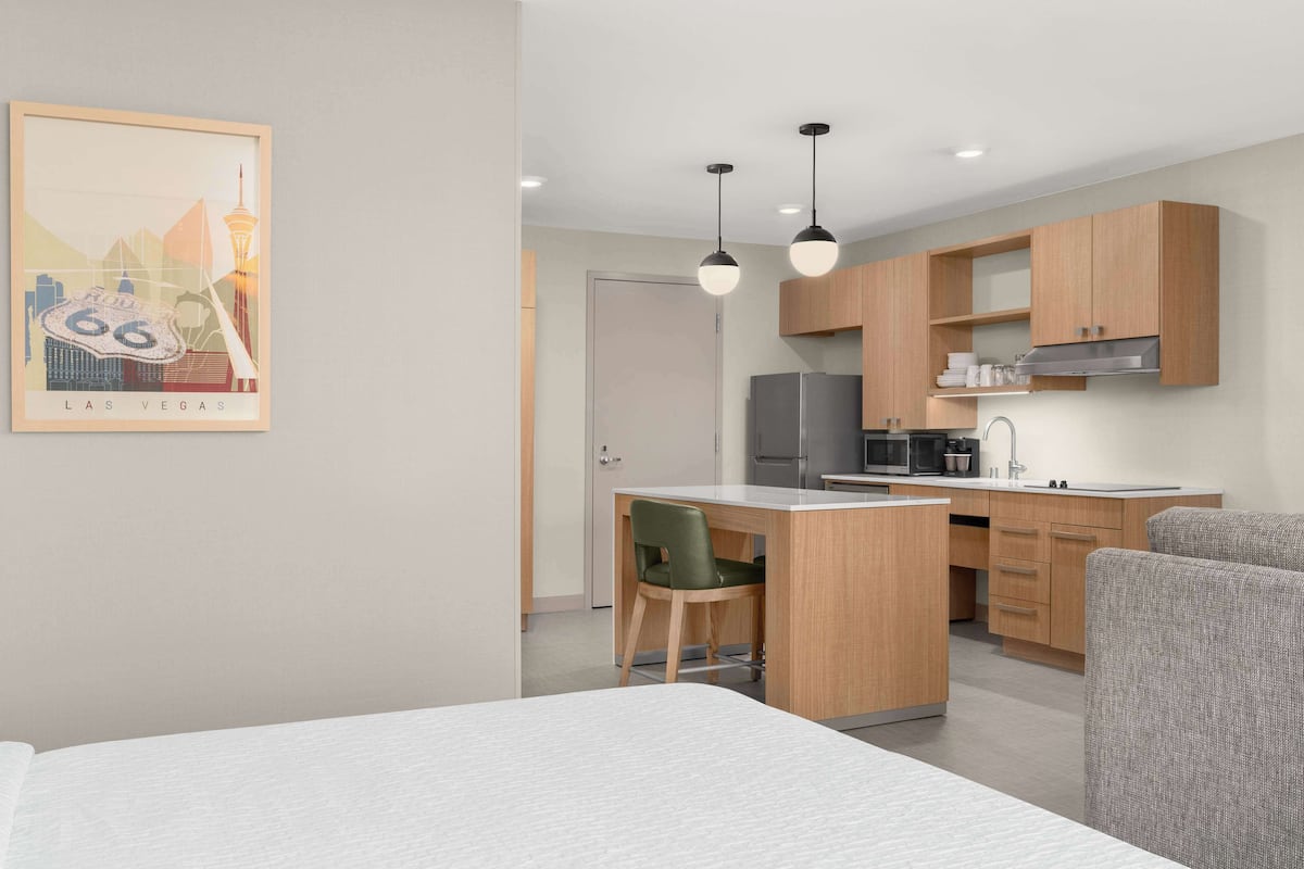 A modern hotel room with a bed, a desk, a chair, a kitchenette, and a framed picture on the wall.