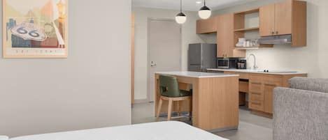 A modern hotel room with a bed, a desk, a chair, a kitchenette, and a framed picture on the wall.