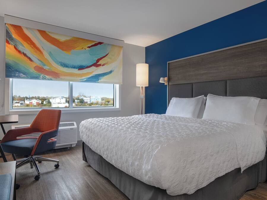 A hotel room with a large bed, a desk, a chair, a colorful abstract painting, a window with a view, and a blue accent wall.