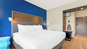 Quarto de hotel moderno com uma cama grande, mesa de cabeceira, luminária fixada na parede e porta que dá para o banheiro.