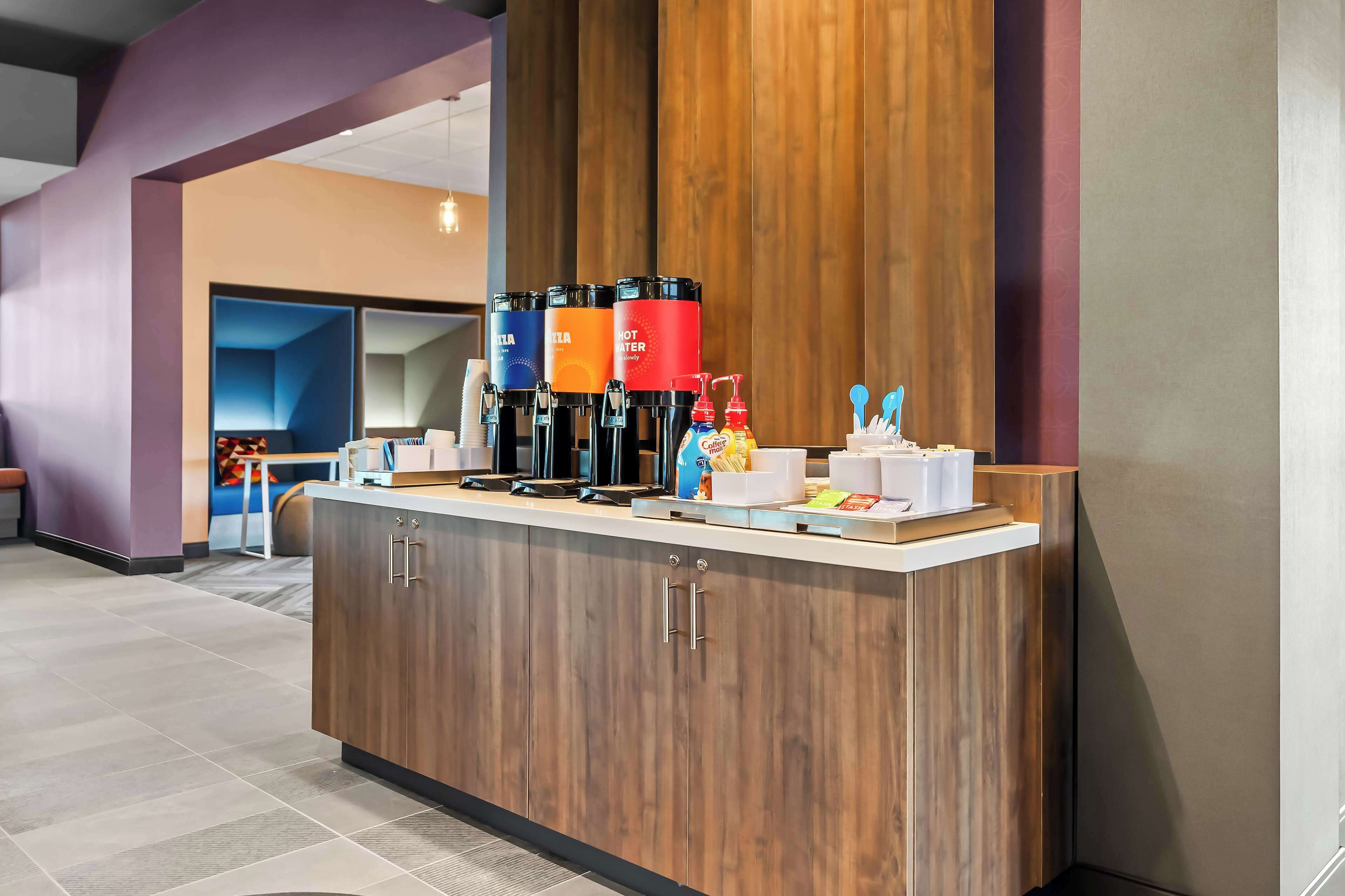Una moderna zona de recepción con mostrador de madera, dispensadores de café y un área de descanso al fondo.