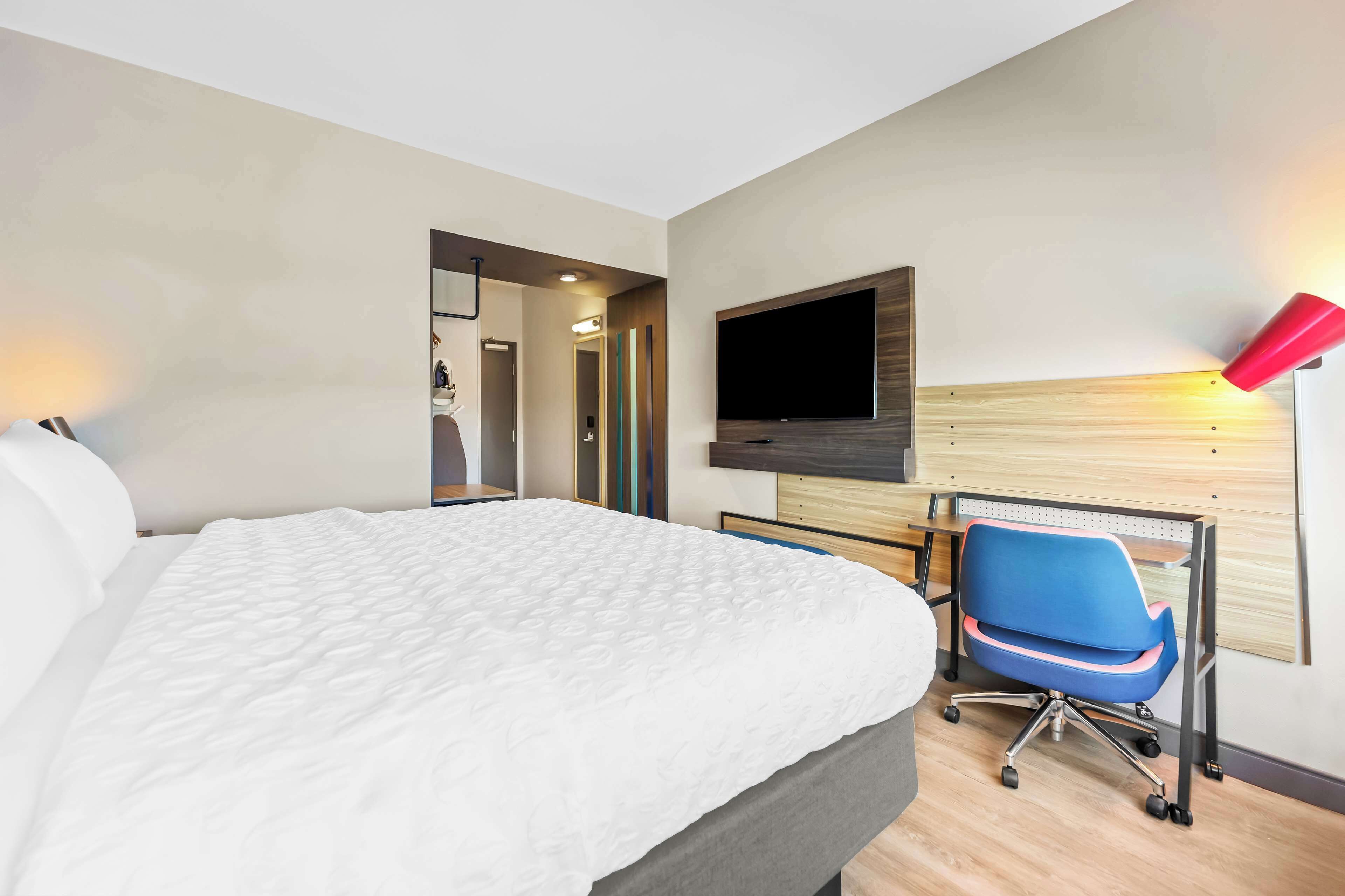 A hotel room with a large bed, a desk with a chair, a TV mounted on the wall, and a red lamp.