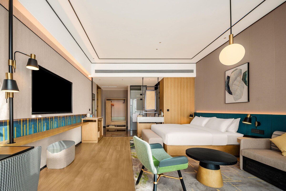 Una habitación de hotel moderna con una cama grande, un escritorio con televisor de pantalla plana, un área de descanso con sofá y sillones, y una mesa de comedor.