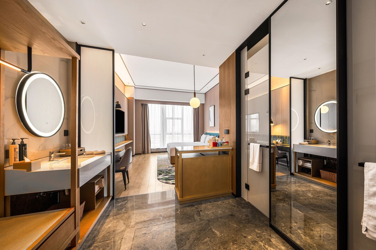 Una habitación de hotel moderna con cama, escritorio, baño con espejo y ventana grande.