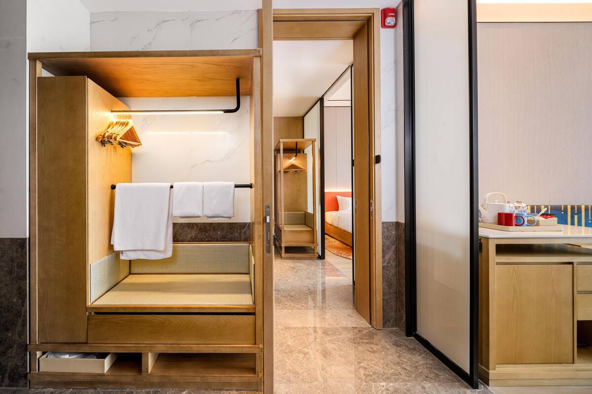 Hotelkamer met een houten bankje, een kleine tafel met lamp, een bed met rode hoofdeinde, en een badkamer met spiegel en handdoekhouder.