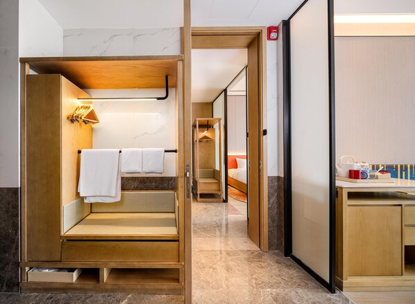 A hotel room with a wooden bench, a small table with a lamp, a bed with a red headboard, and a bathroom with a mirror and towel rack. - Hilton Garden Inn Fenyang Yulongwan (Lvliang)