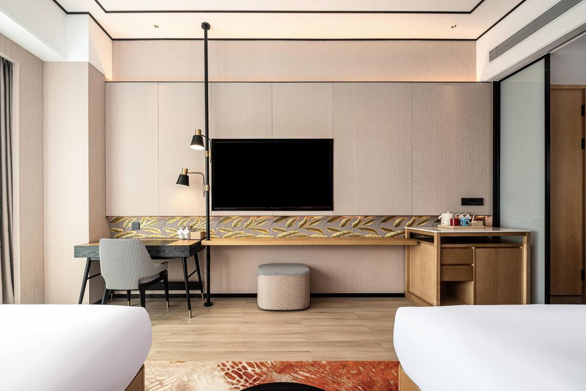 Een moderne hotelkamer met een flatscreen televisie aan de muur, een bureau met stoel, een klein voetenbankje en twee bedden.