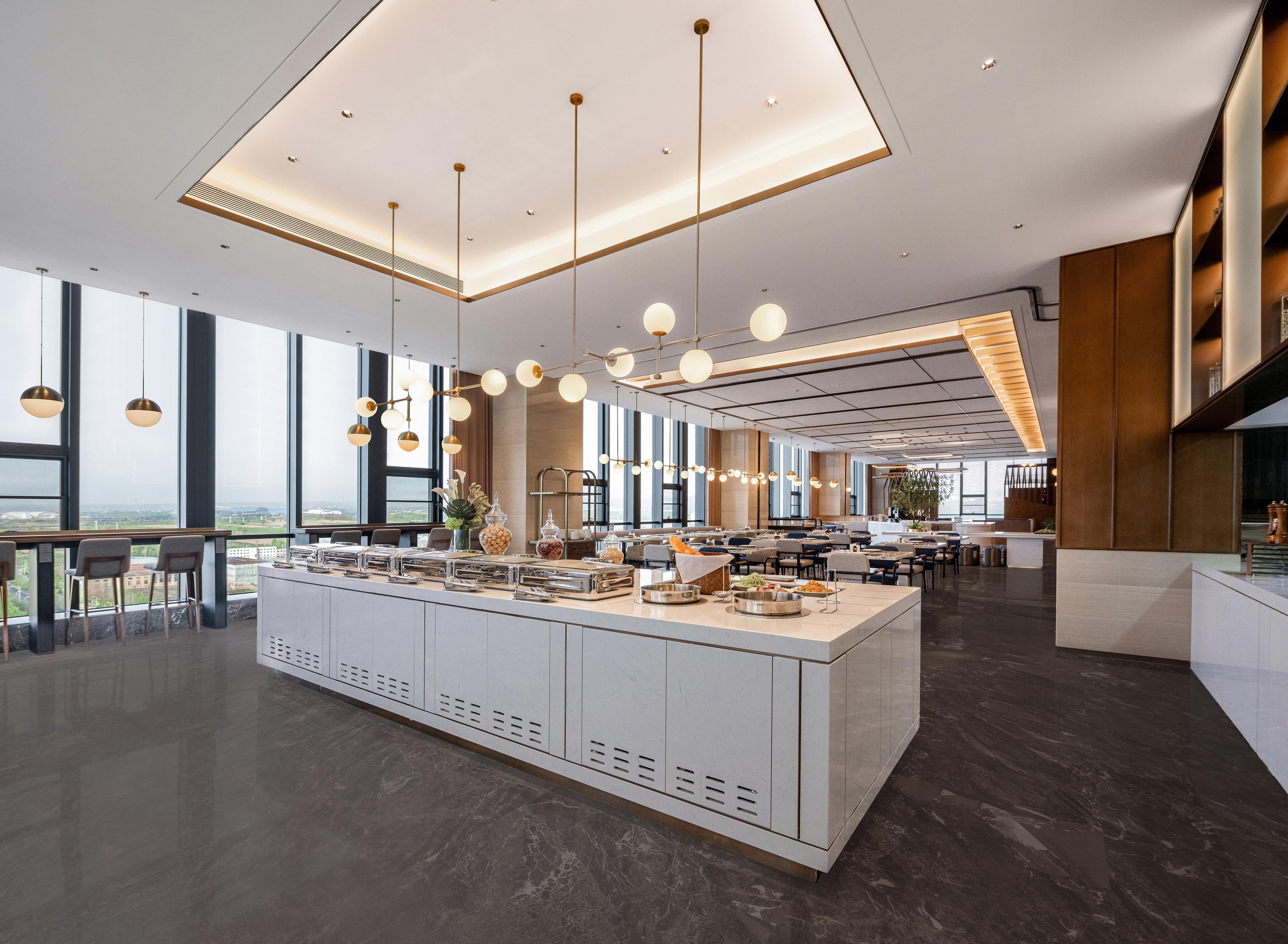 Un comedor de hotel moderno con una isla central, iluminación colgante y amplios ventanales con vistas.