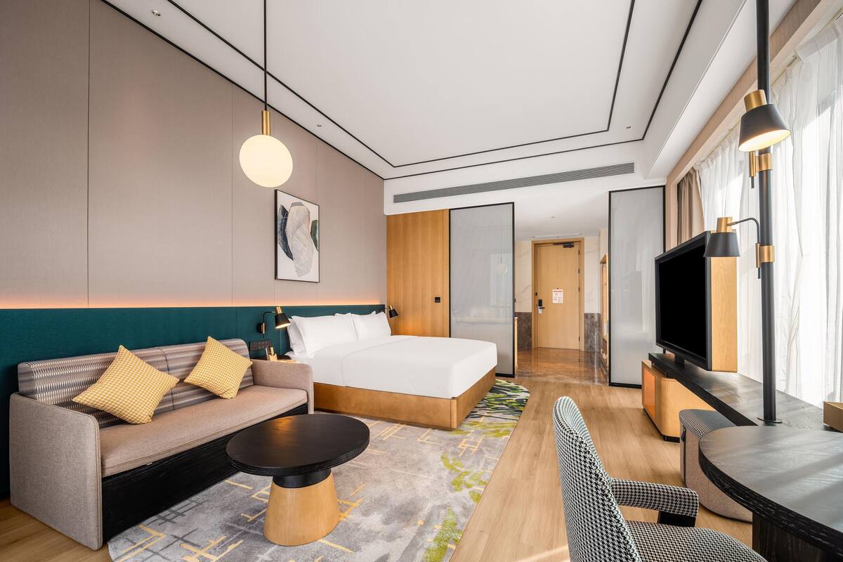 Een moderne hotelkamer met een bed, bank, salontafel, televisie en uitzicht op de gang.