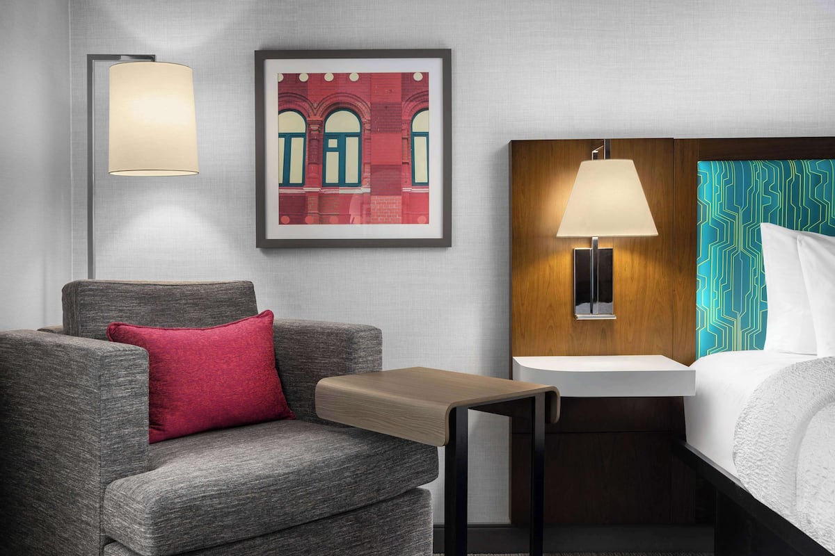 Et hotelværelse med en grå sofa, et lille træsidebord, en seng med hvide sengetøj, en vægmonteret lampe og et indrammet billede af en bygning.