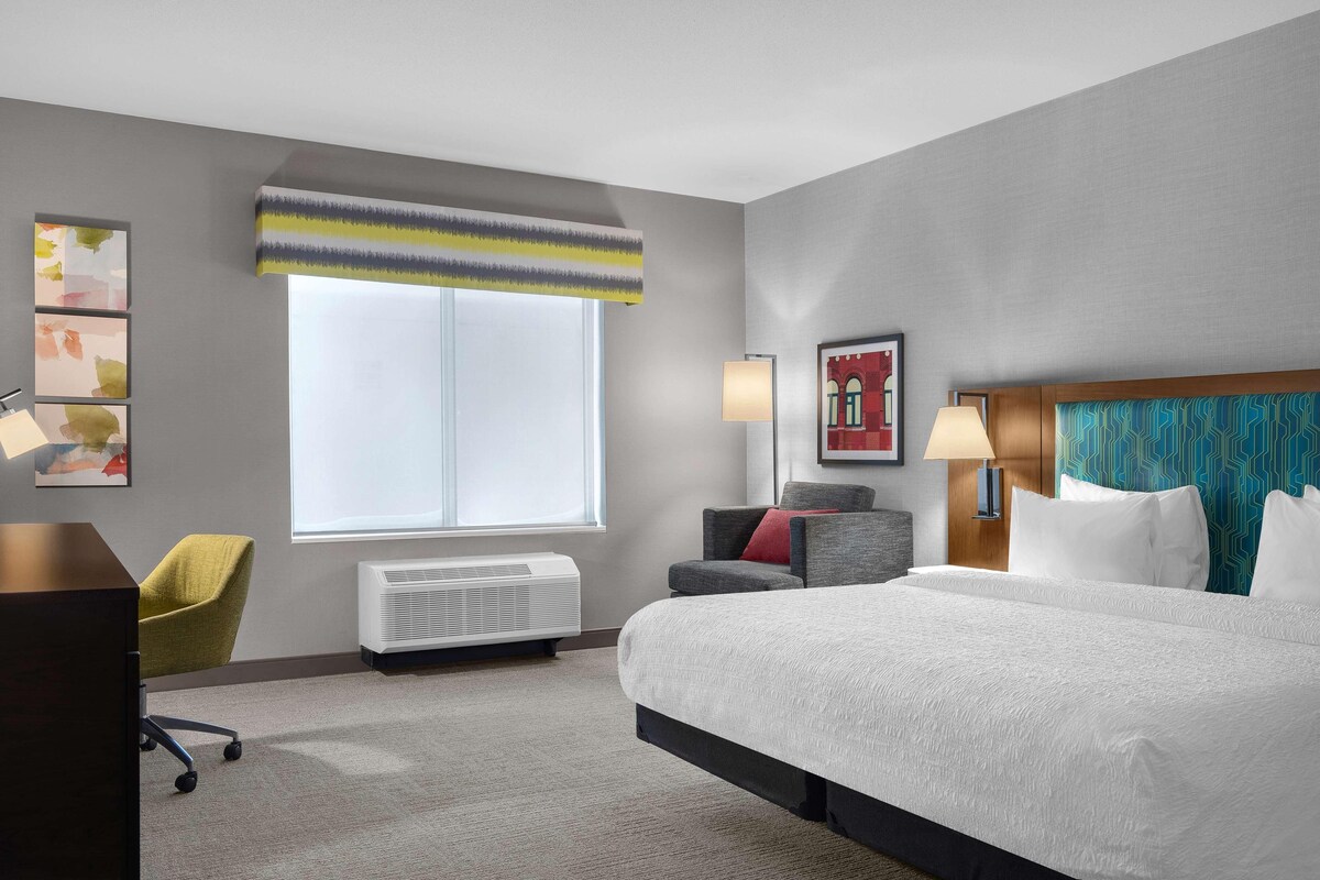 A hotel room with a large bed, a desk, a chair, a window with a view, and a wall with artwork.
