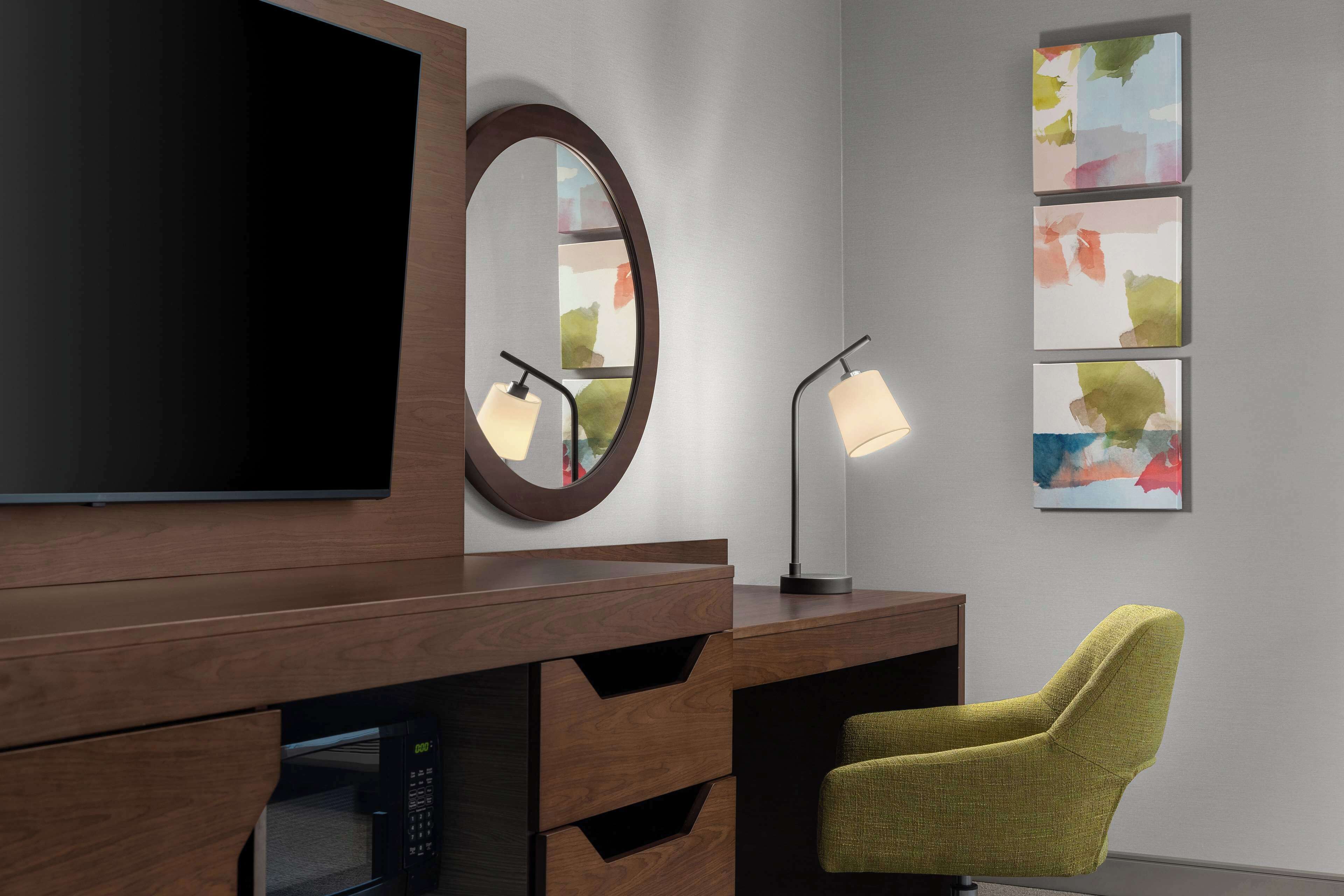 Et moderne hotelværelse med et træskrivebord, et fladskærms-tv, et rundt spejl, en skrivebordslampe, en grøn stol og abstrakt kunst på væggen.
