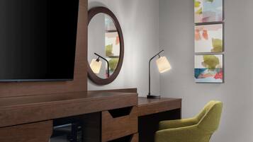 Et moderne hotelværelse med et træskrivebord, et fladskærms-tv, et rundt spejl, en skrivebordslampe, en grøn stol og abstrakt kunst på væggen.