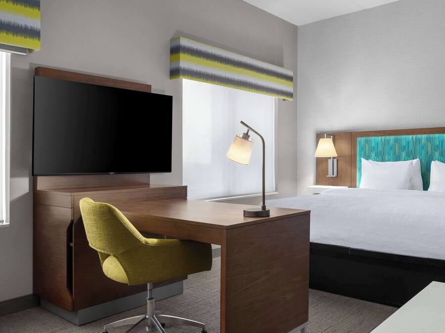 Ein Hotelzimmer mit einem Holztisch, einem Fernseher, einem Bett mit weißer Bettwäsche und einem Fenster mit Vorhängen.