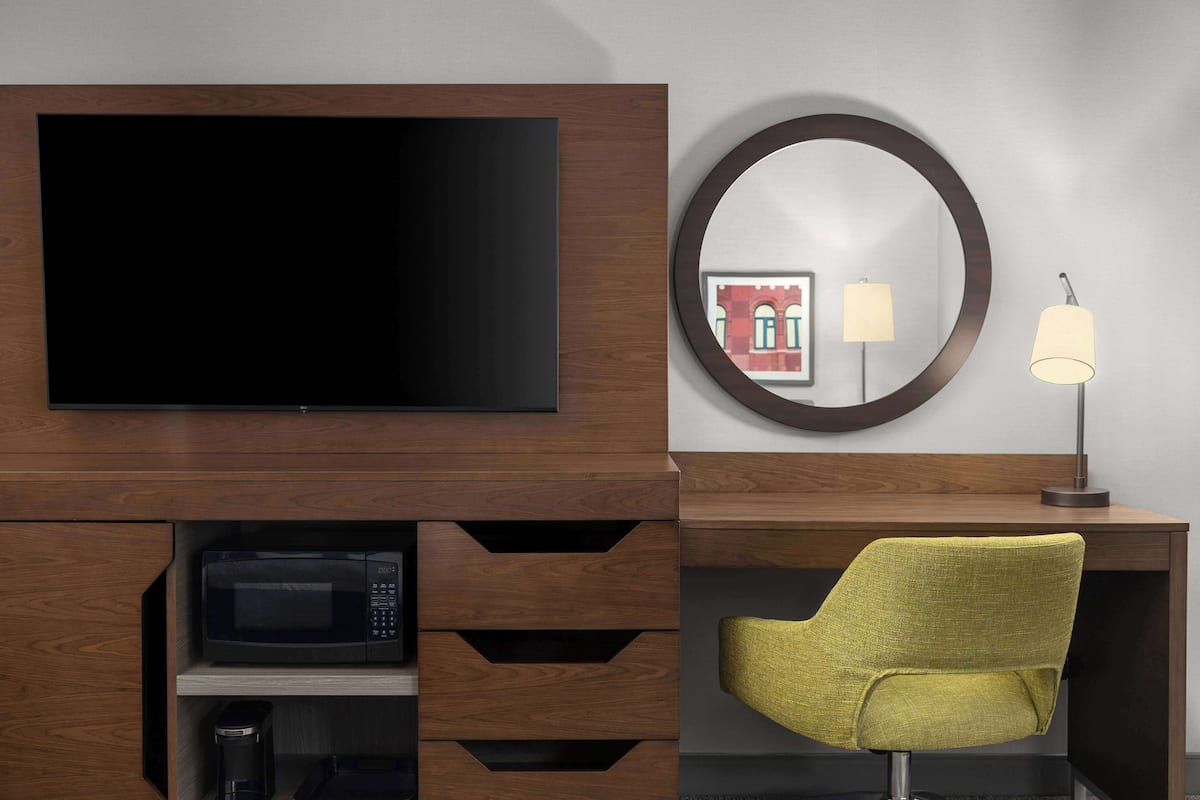 Et moderne værelse med et fladskærms-TV monteret på et træskab, et rundt spejl på væggen, en lampe, en mikrobølgeovn og en grøn stol.