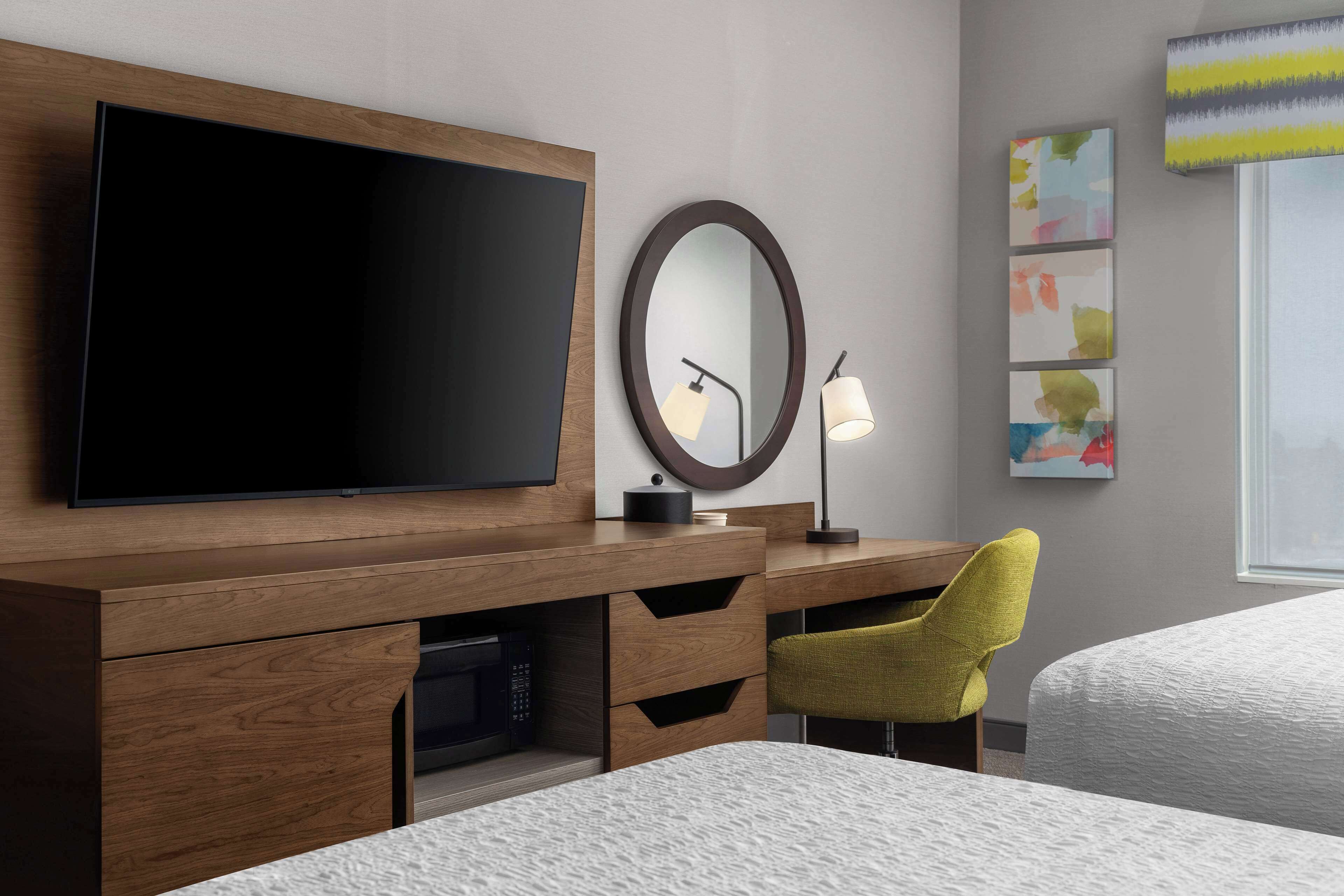 A hotel room with a wooden desk, a flat-screen TV, a round mirror, a lamp, and a bed.