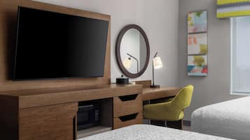A hotel room with a wooden desk, a flat-screen TV, a round mirror, a lamp, and a bed.