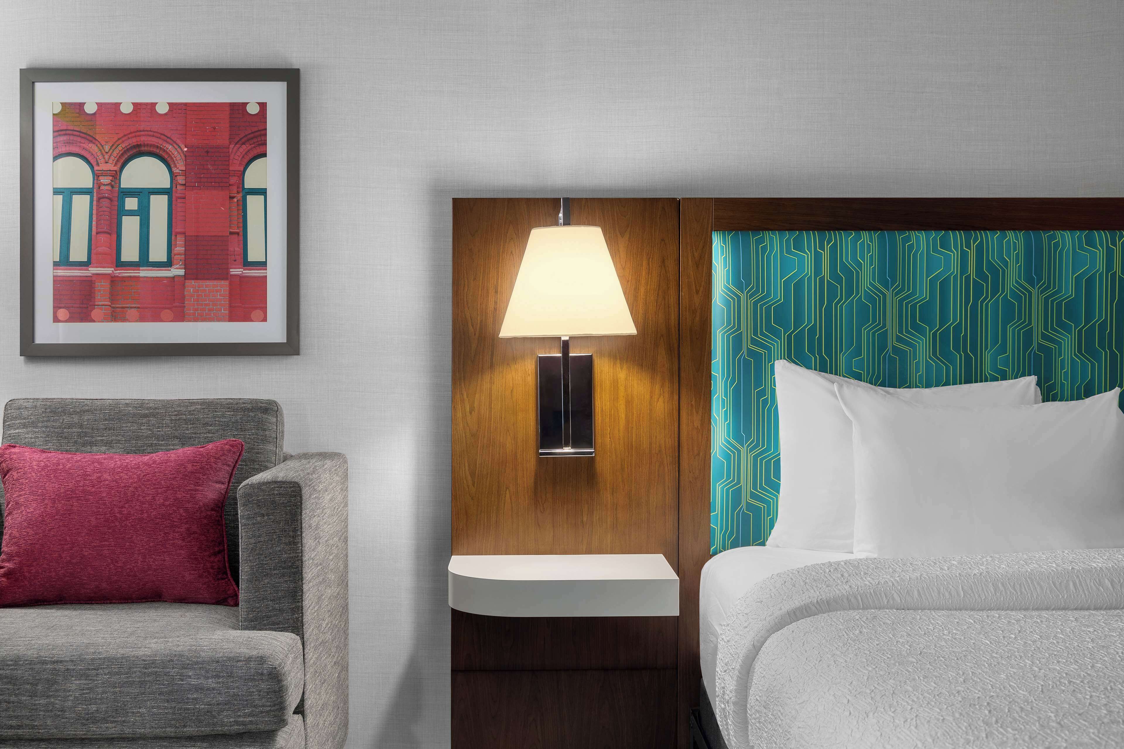 Habitación de hotel con sofá gris, almohada roja, lámpara de pared y cama con cabecera estampada.