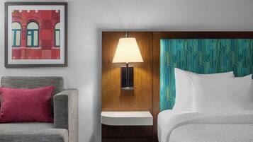 Habitación de hotel con sofá gris, almohada roja, lámpara de pared y cama con cabecera estampada.