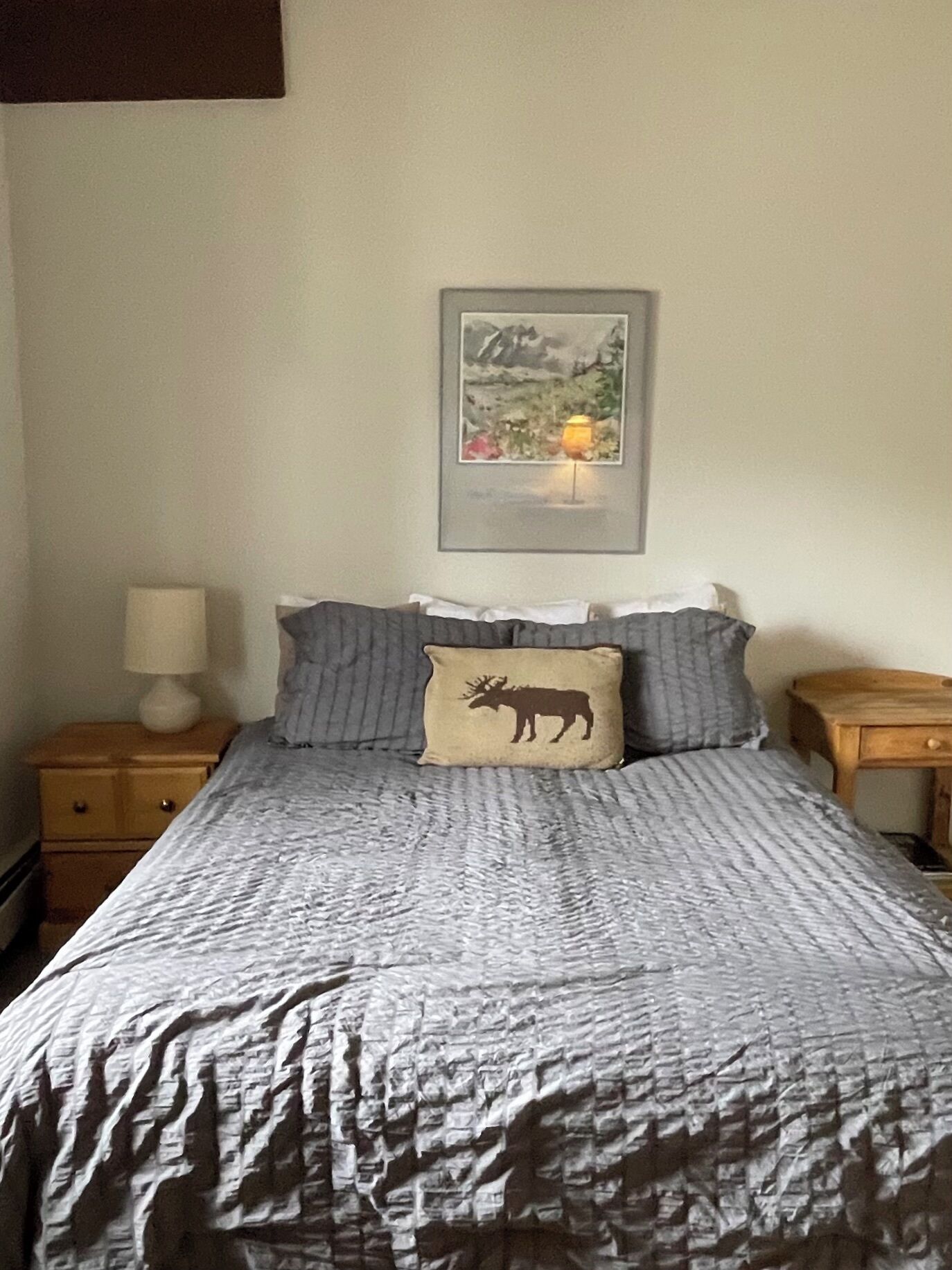 Ein Bett mit grau-weiß gestreifter Bettdecke, einem Kopfkissen mit Elch-Motiv und einem Landschaftsbild im Rahmen darüber.
