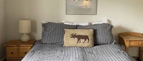 Ein Bett mit grau-weiß gestreifter Bettdecke, einem Kopfkissen mit Elch-Motiv und einem Landschaftsbild im Rahmen darüber.