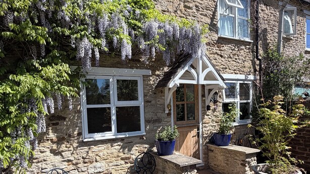 A stone building with a window, a door, and a small porch area with a table and chairs. - 17th-Century Cotswold Cottage with South-Facing Orchard. (BICESTER)