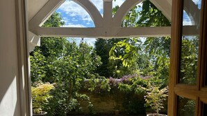 A view through a doorway into a garden with a stone pathway, potted plants, and a variety of trees and shrubs. - 17th-Century Cotswold Cottage with South-Facing Orchard. (BICESTER)
