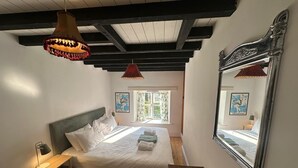 A bedroom with a large bed, a wooden floor, a nightstand, a lamp, a mirror, and a window with curtains. - 17th-Century Cotswold Cottage with South-Facing Orchard. (BICESTER)