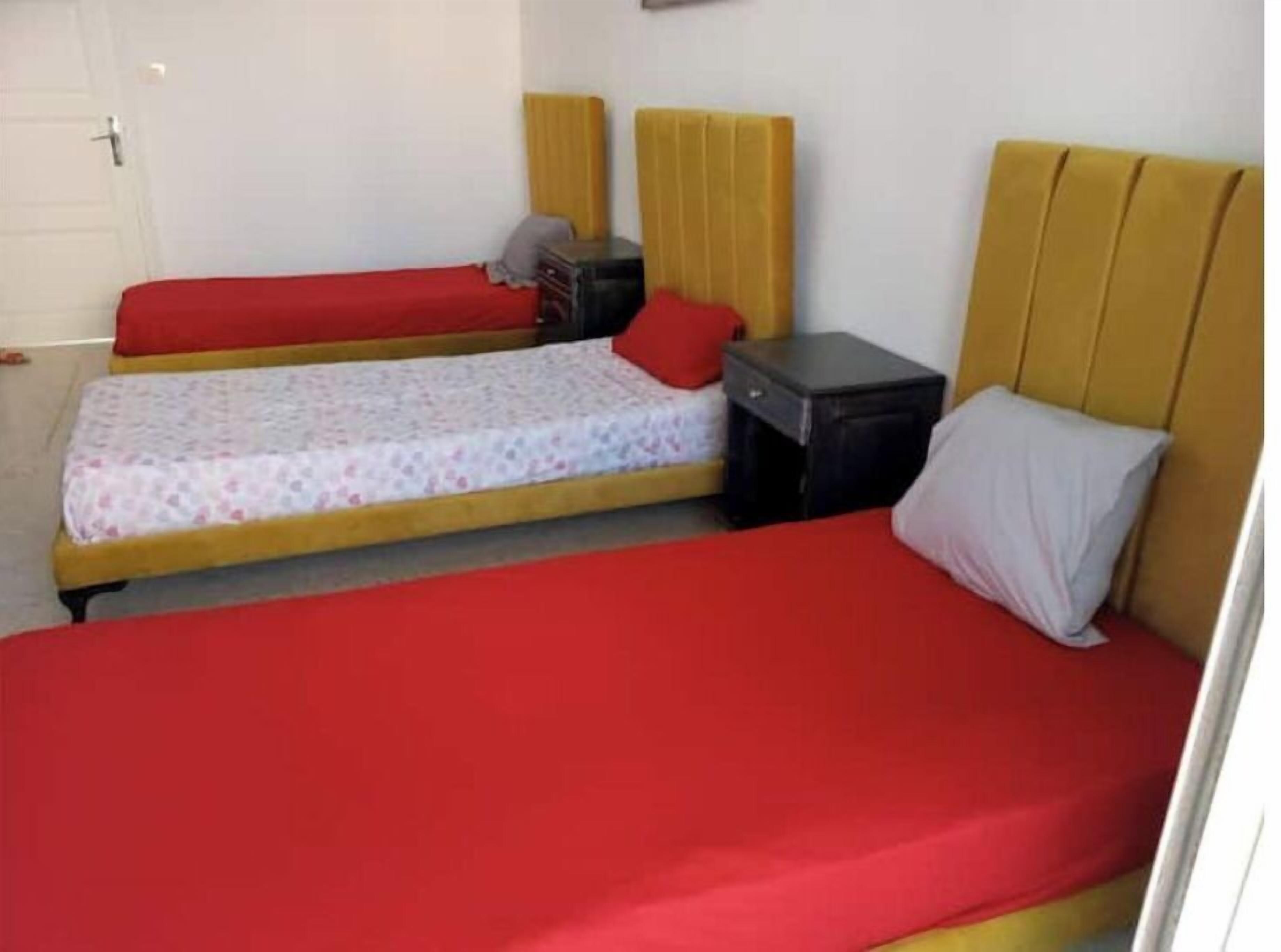 Una habitación con dos camas individuales, cada una con una colcha roja y una almohada. Hay una mesita de noche negra pequeña con una lámpara entre las camas.