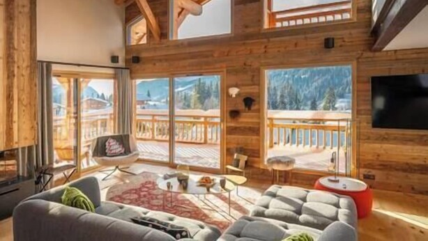 A spacious living room with a large gray sofa, a red ottoman, and a wooden ceiling. There are large windows offering a view of a snowy mountain landscape. - 5 bedroom Luxury chalet with panoramic view, nordic bath, high quality finis (Les Gets)