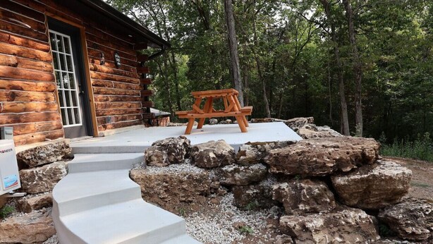 Una cabaña de troncos con una escalera de piedra que lleva a una mesa de picnic de madera en un área boscosa.
