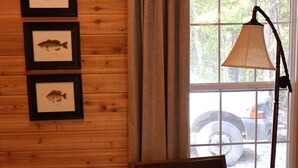 Un interior de madera con una silla, cuadros de peces enmarcados, una ventana con cortinas y una lámpara.