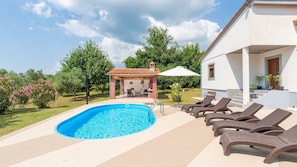 Una piscina con sillones de descanso, un porche cubierto y un jardín con árboles y flores.