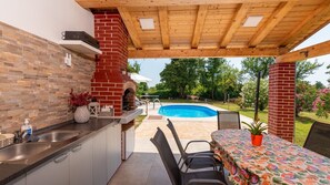 Una cocina exterior con horno de ladrillo, piscina y mesa con sillas.
