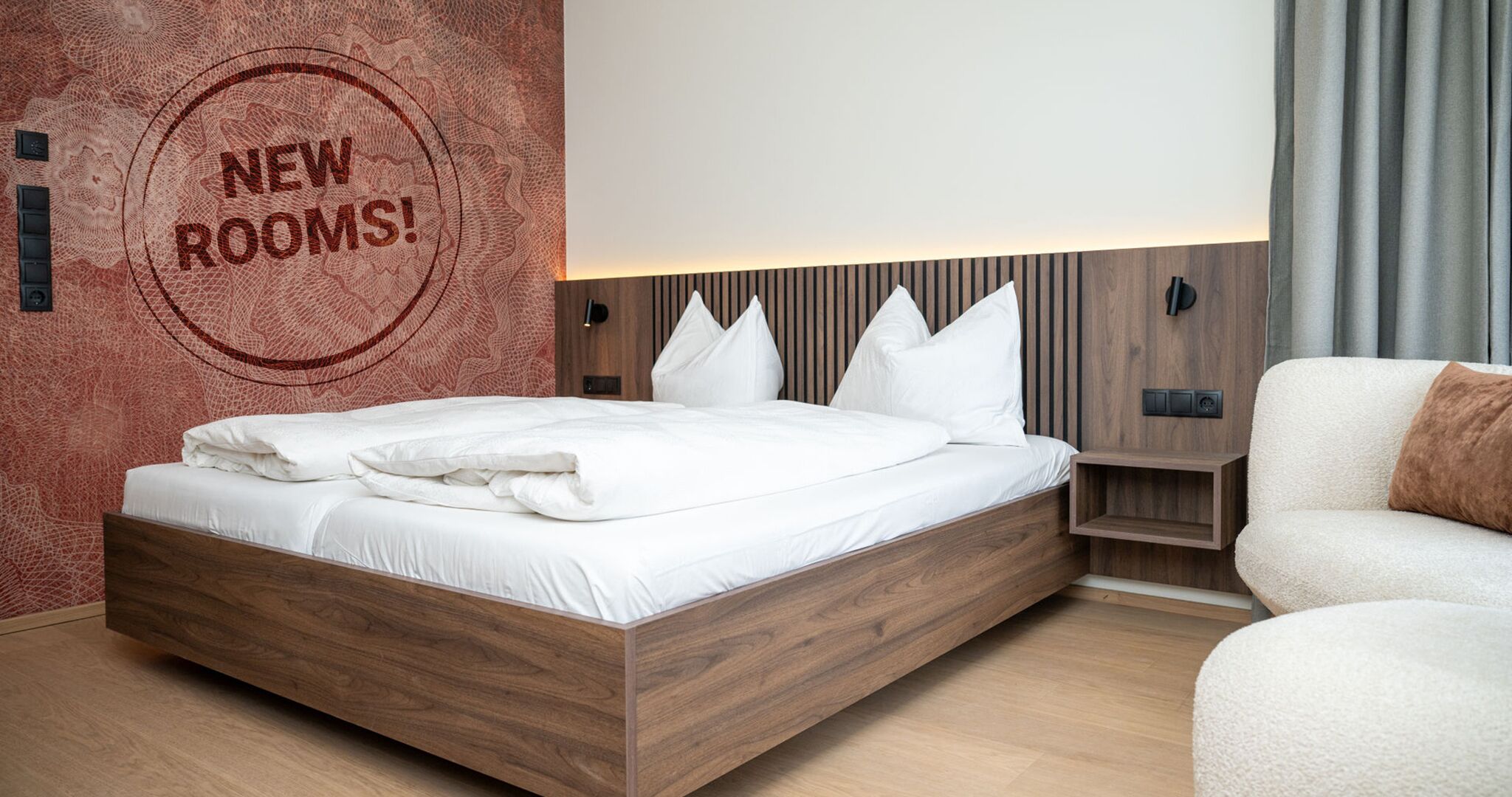 A modern bedroom with a wooden bed, white bedding, and a wall with a 'NEW ROOMS!' sign.