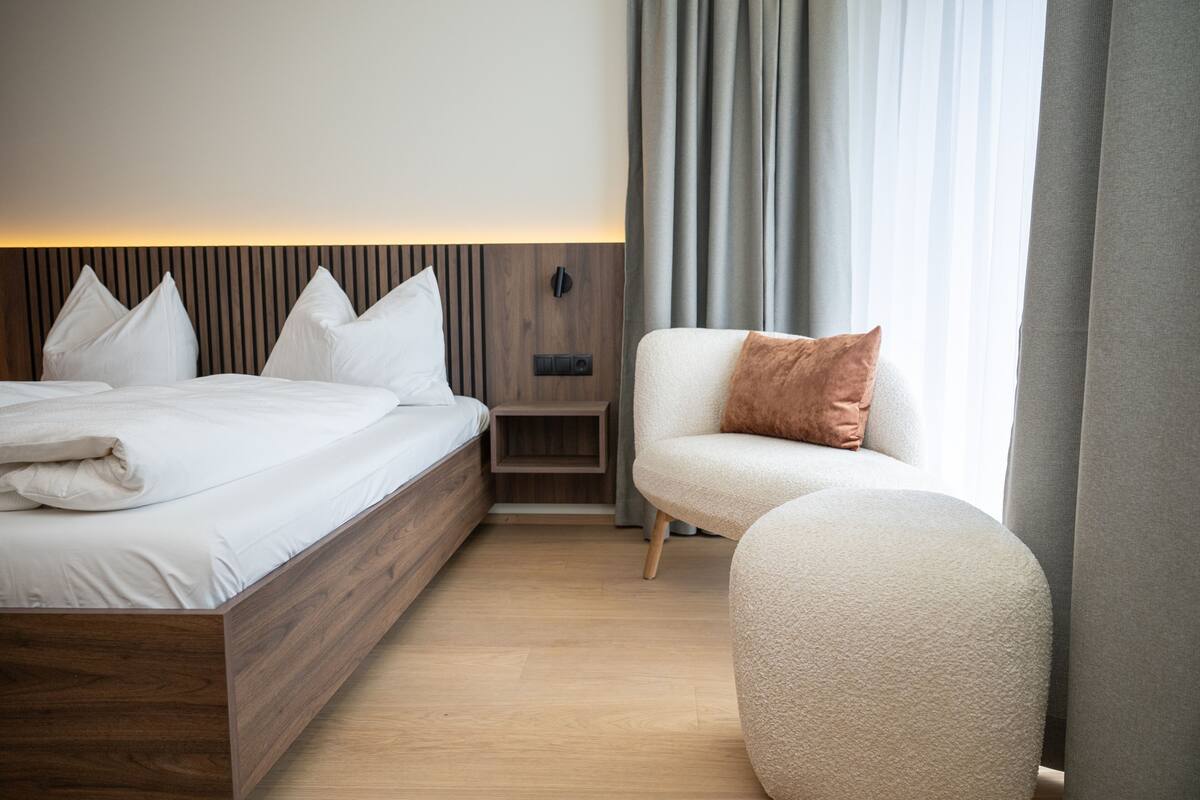 Ein modernes Hotelzimmer mit einem hölzernen Bett, einem kleinen Nachttisch, einem Sessel und einem großen Fenster mit Vorhängen.