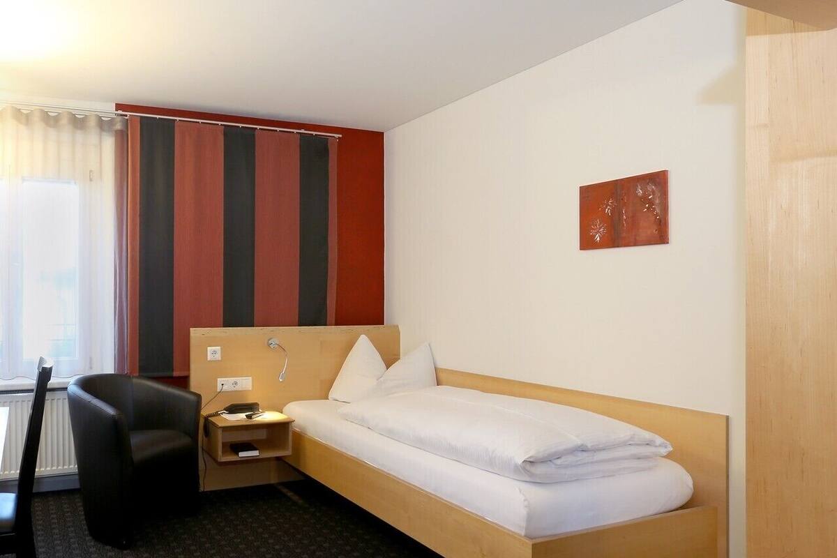 Ein Hotelzimmer mit einem Einzelbett, einem Schreibtisch, einem Stuhl und einem Fenster mit Vorhängen.