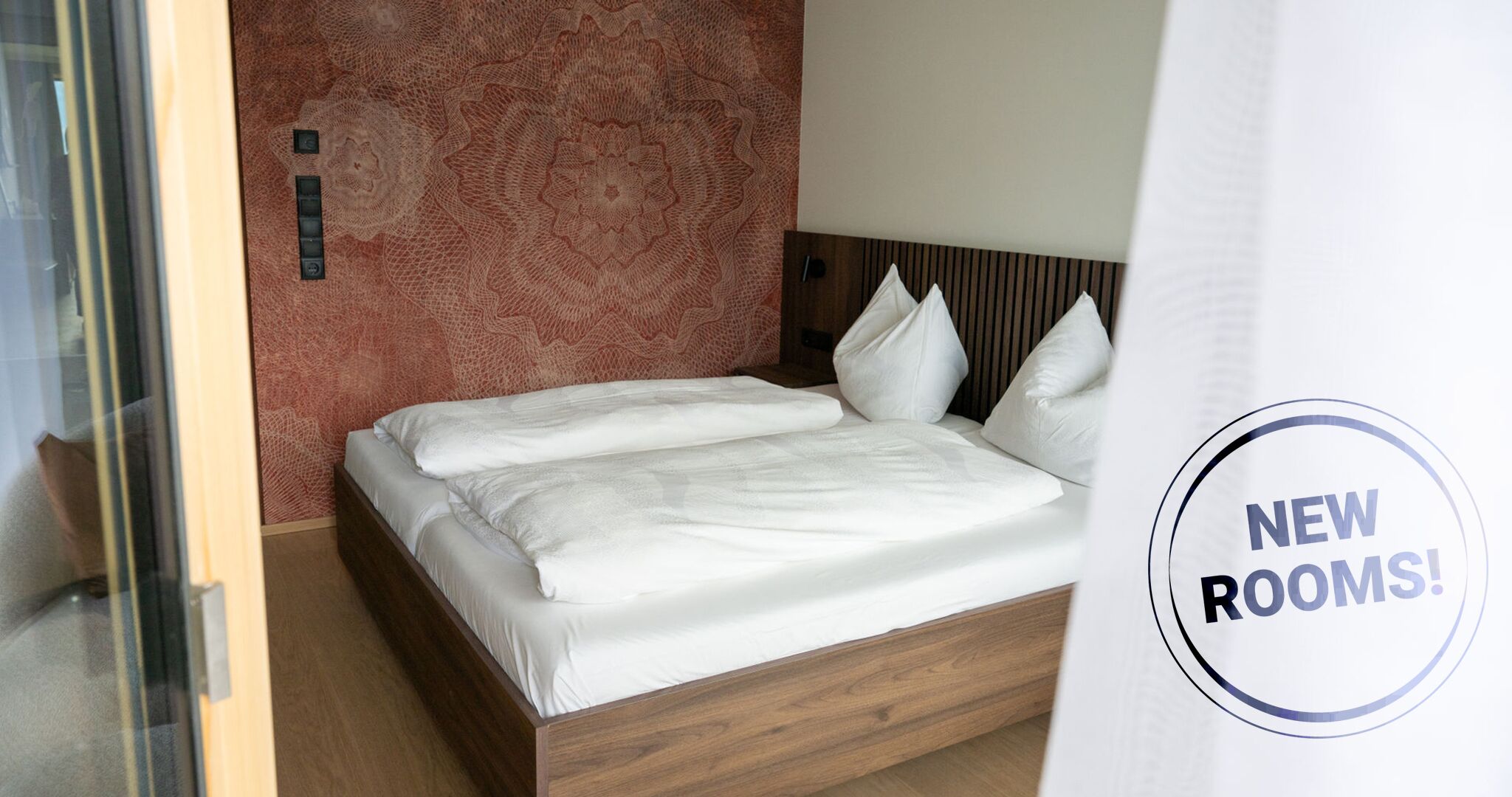 A neatly made bed with white linens in a room with a patterned wall and a wooden headboard.