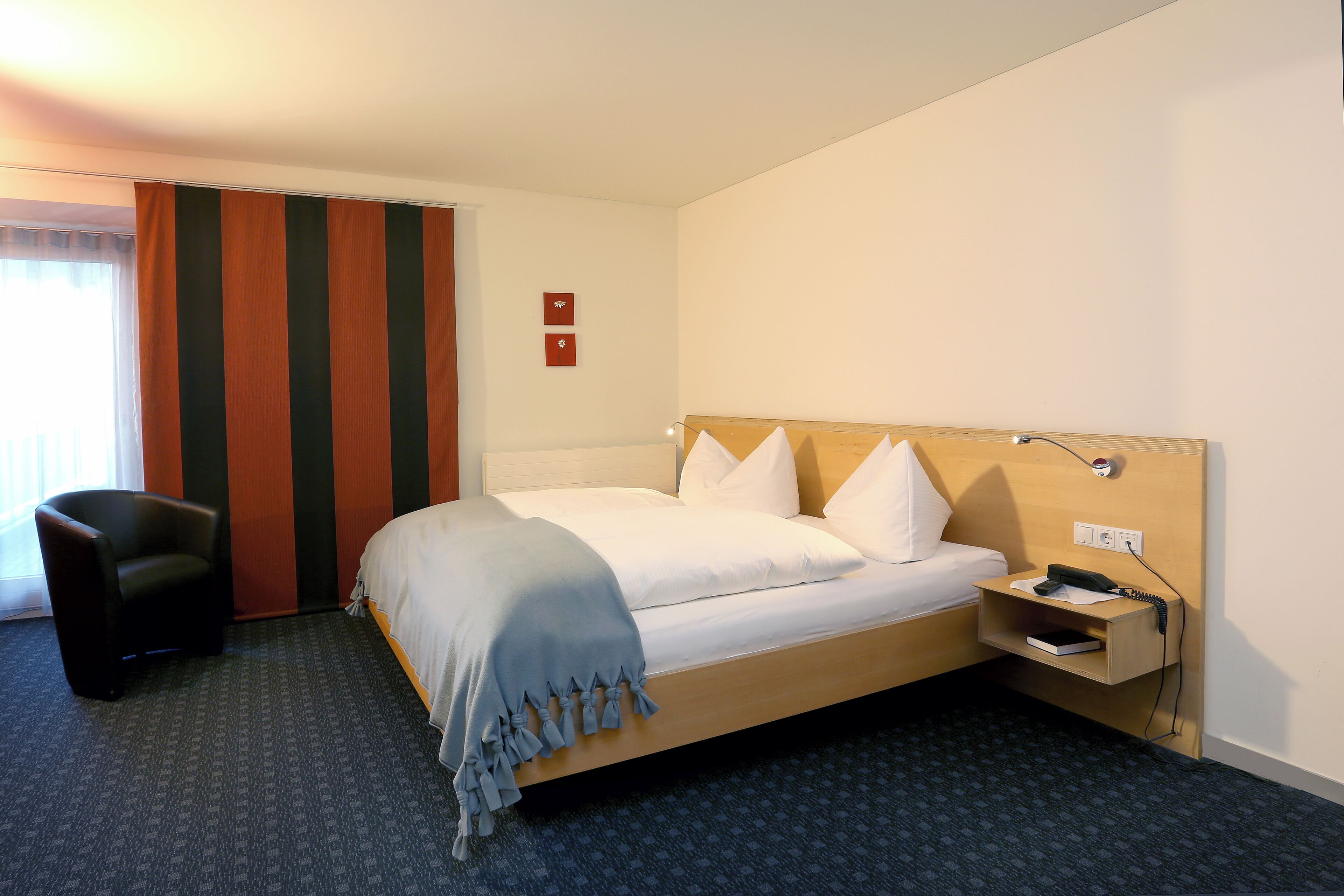 Una habitación de hotel con una cama individual, una mesita de noche con un teléfono y un libro, una silla y una cortina a rayas.