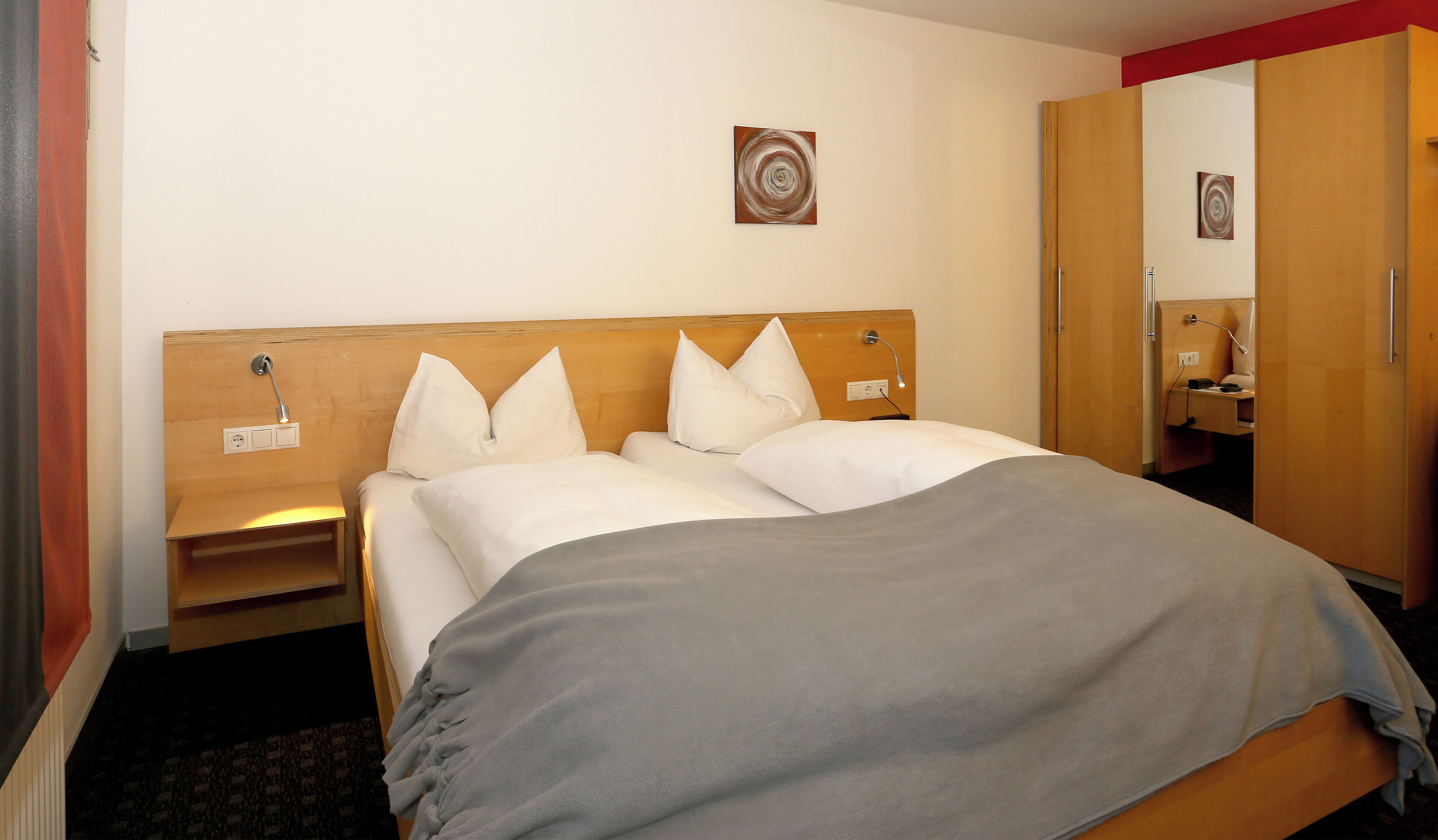 A hotel room with a wooden headboard, a bed with white linens, a gray blanket, a nightstand with a lamp, and a wooden wardrobe.