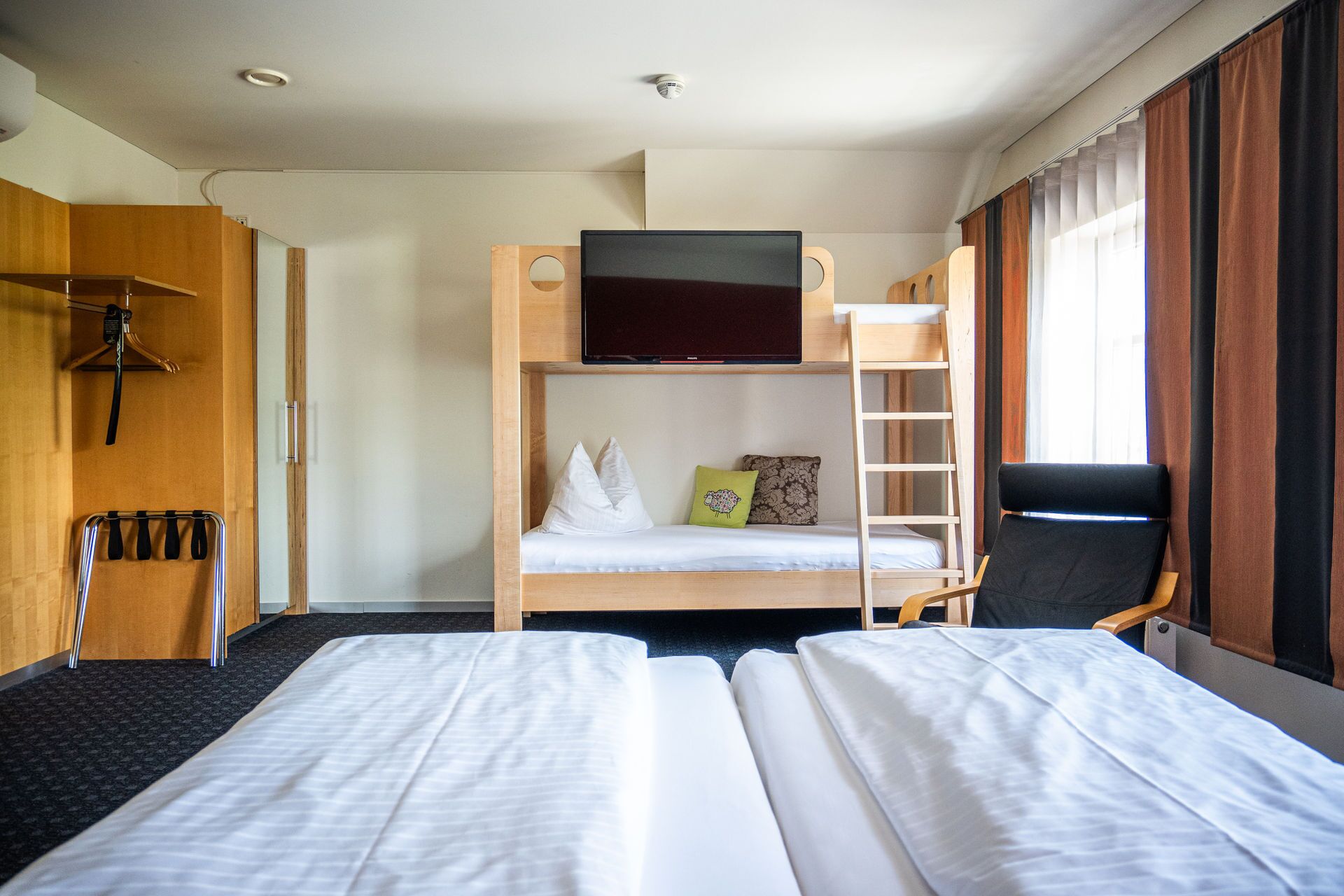 A hotel room with a bunk bed, a desk with a chair, a TV mounted above the bed, and a window with curtains.