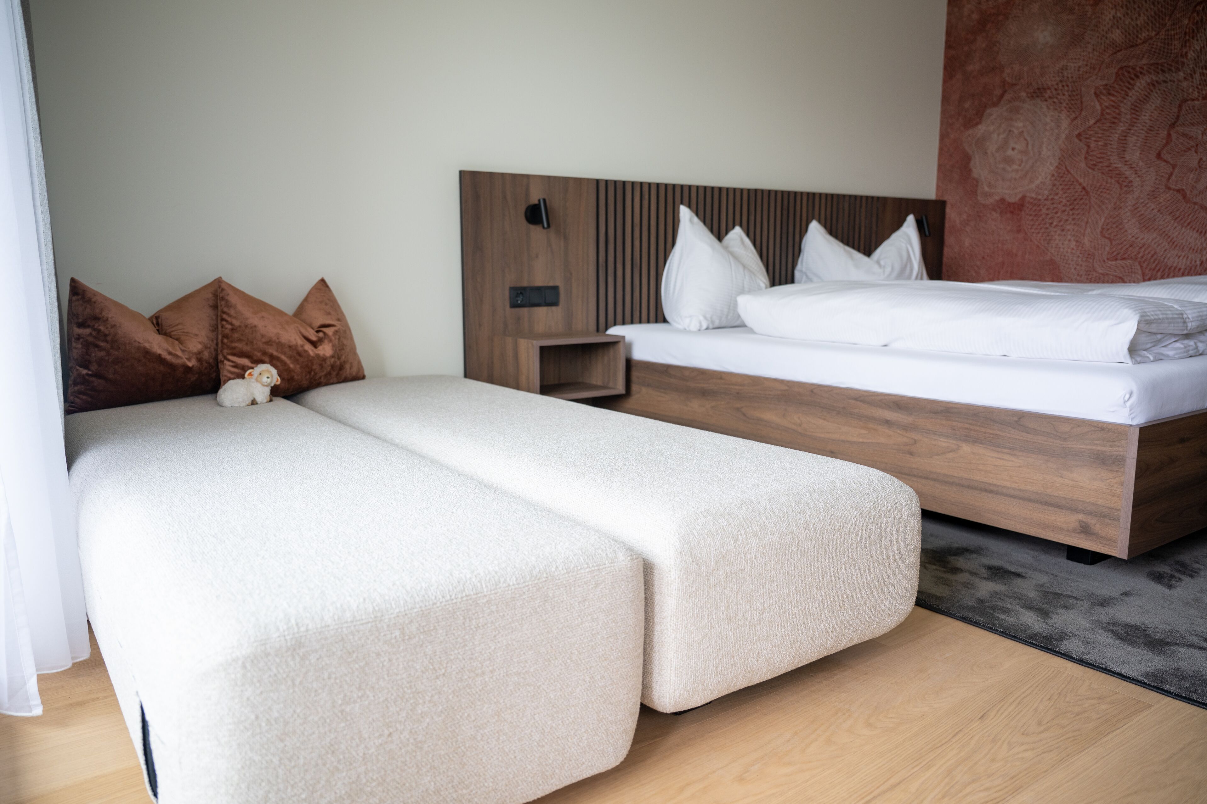 A modern hotel room with a wooden headboard, a large bed, a smaller sofa bed, and a nightstand.