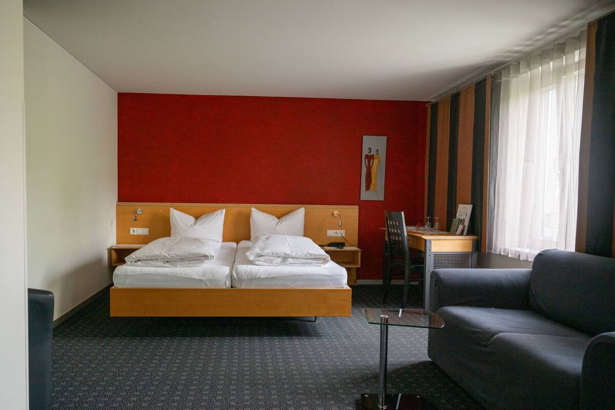 Ein Hotelzimmer mit einem Doppelbett, einem Schreibtisch, einem Stuhl und einem großen Fenster mit Vorhängen.