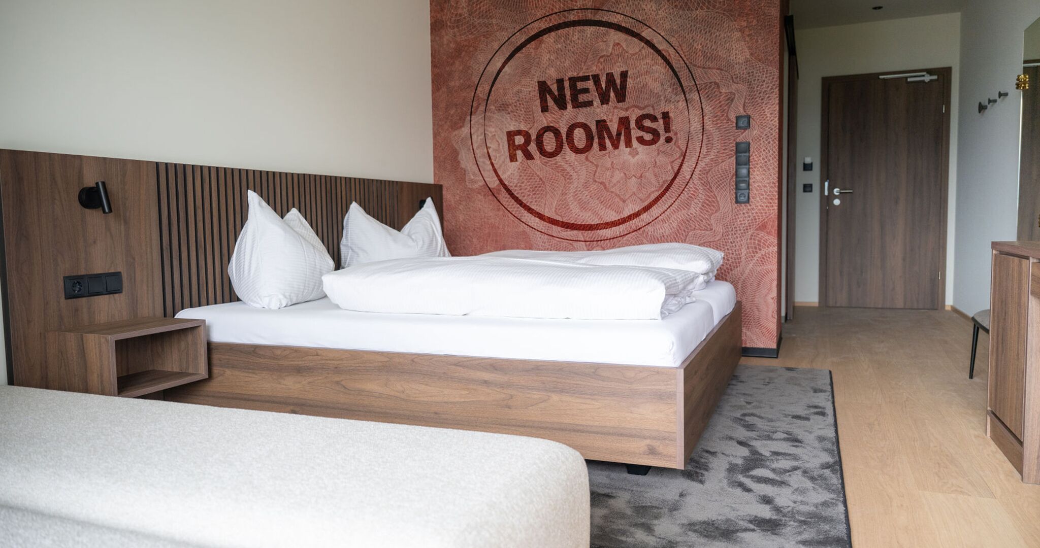 Een moderne hotelkamer met een houten hoofdbord, een groot bed met witte beddengoed, een nachtkastje en een muur met een 'NEW ROOMS!' bord.