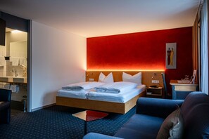 Una habitación de hotel con cama, una mesita pequeña, un sofá, una mesita de noche y un baño visible a través de una puerta abierta.
