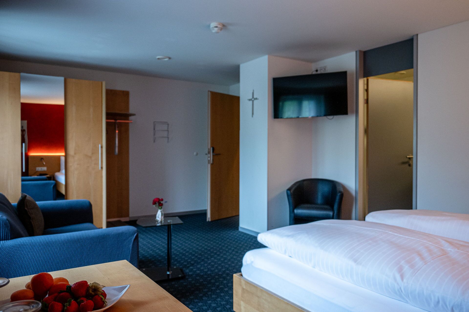 Habitación de hotel con cama, escritorio con fruta, una silla, televisor y una cruz en la pared.