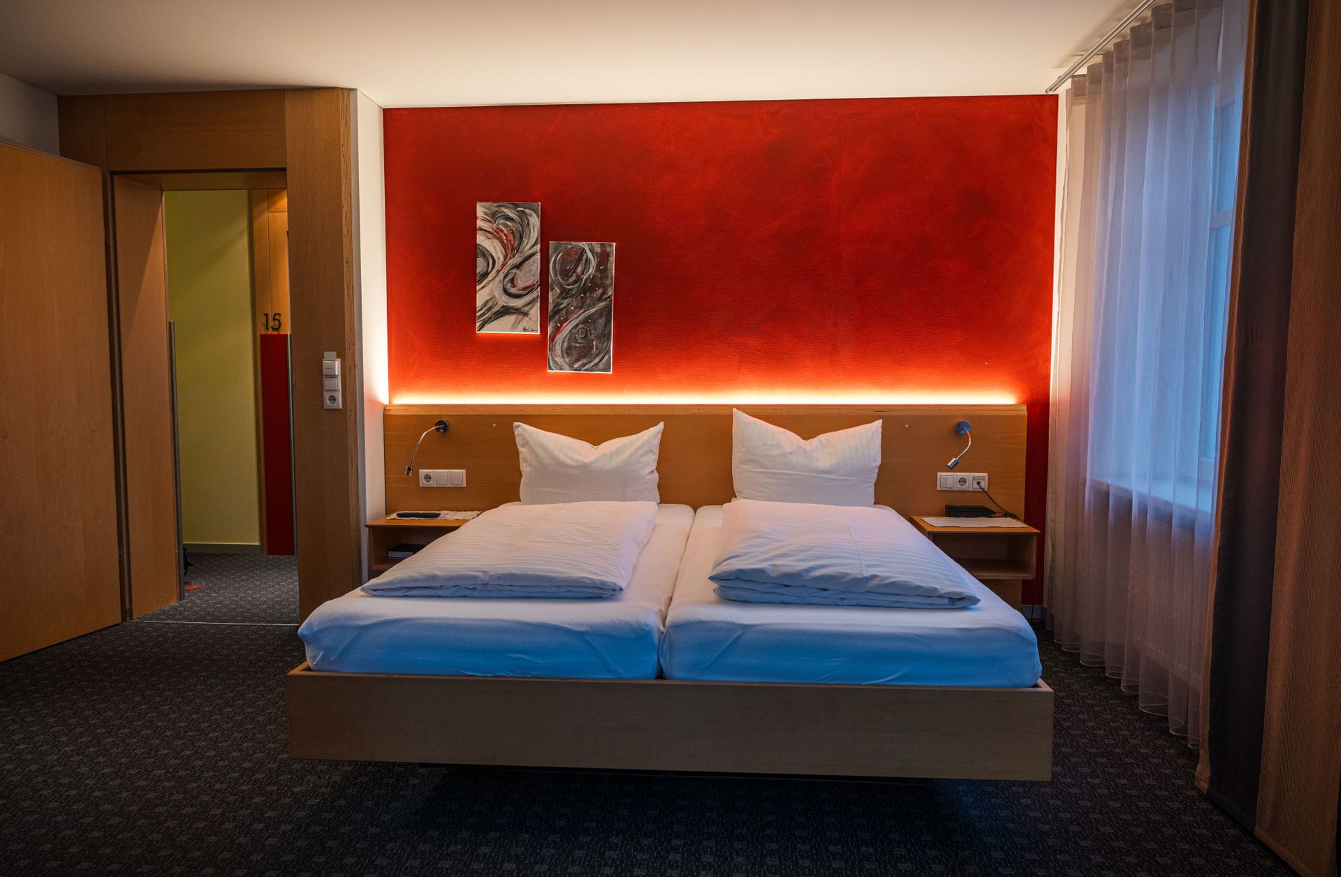 Cama doble con sábanas blancas, pared de acento roja, dos mesitas de noche con lámparas, y una puerta con el número 15.