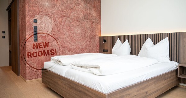 Un dormitorio moderno con estructura de cama de madera, ropa de cama blanca y una pared con estampado rojo texturizado y un letrero que dice 'HABITACIONES NUEVAS!'.