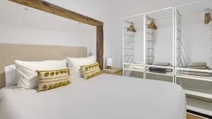 Un dormitorio con una cama grande, ropa de cama blanca, un cabecero de madera, un armario blanco y una mesita de noche pequeña con una lámpara.