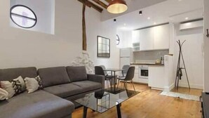 Una sala moderna con un sofá gris, una mesa de centro de vidrio y una zona de comedor con sillas. Se ve una cocina al fondo con armarios blancos y una lámpara colgante.